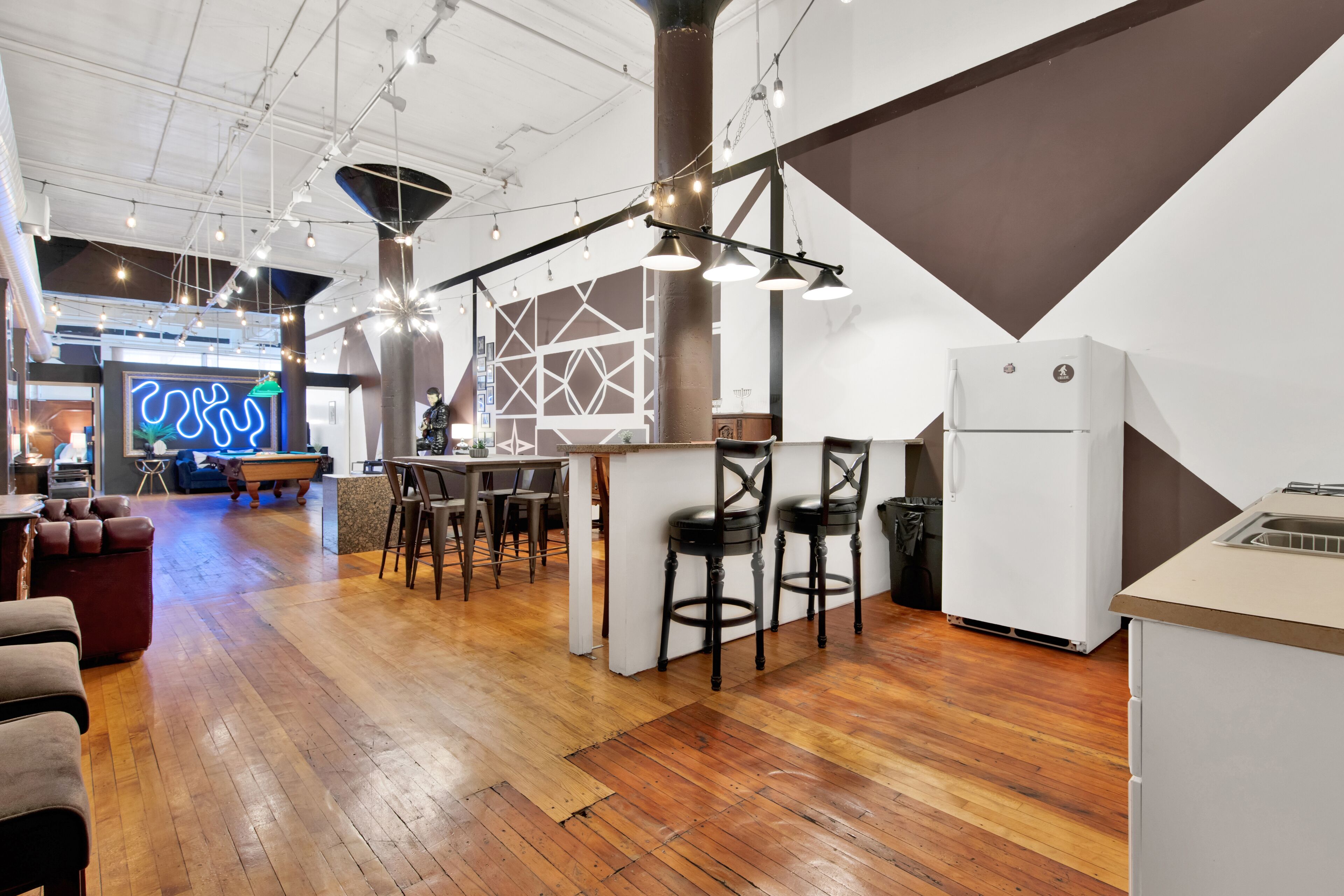 A modern interior with a wooden floor, a bar area with stools, and a neon sign in the background.