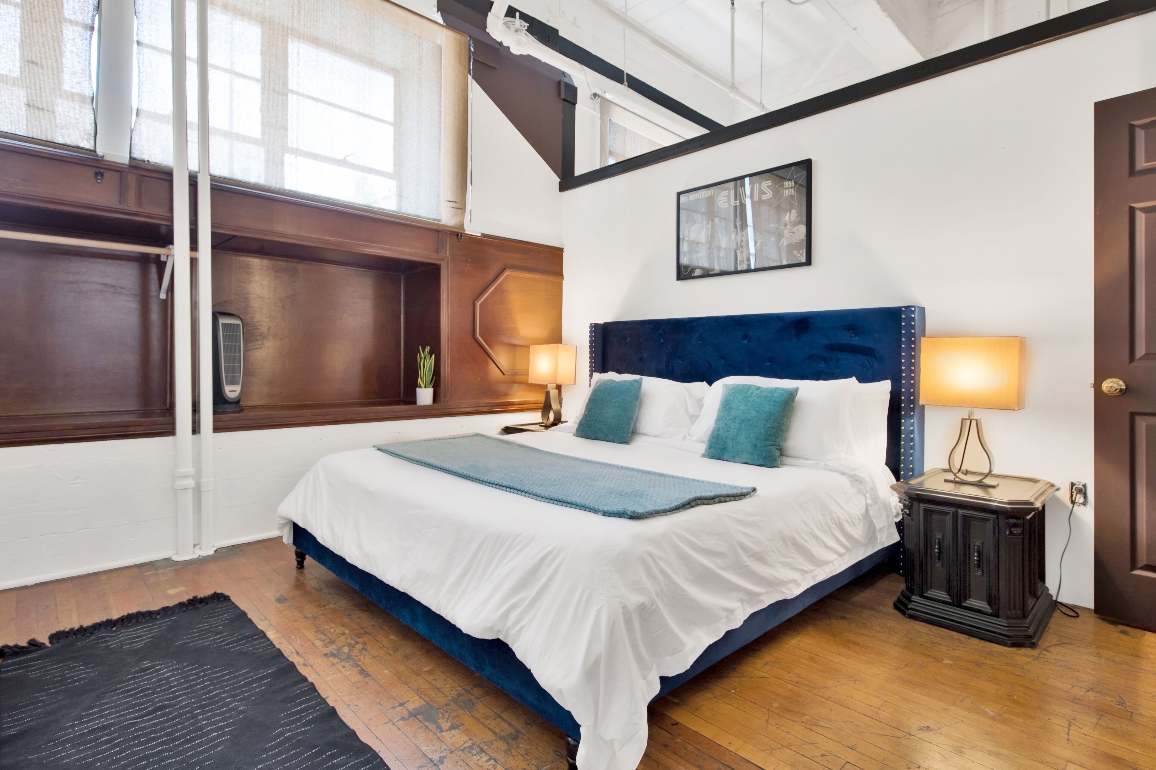 Une chambre avec un grand lit, un plancher en bois, une tête de lit sombre, une table de chevet avec une lampe et une fenêtre avec des rideaux.