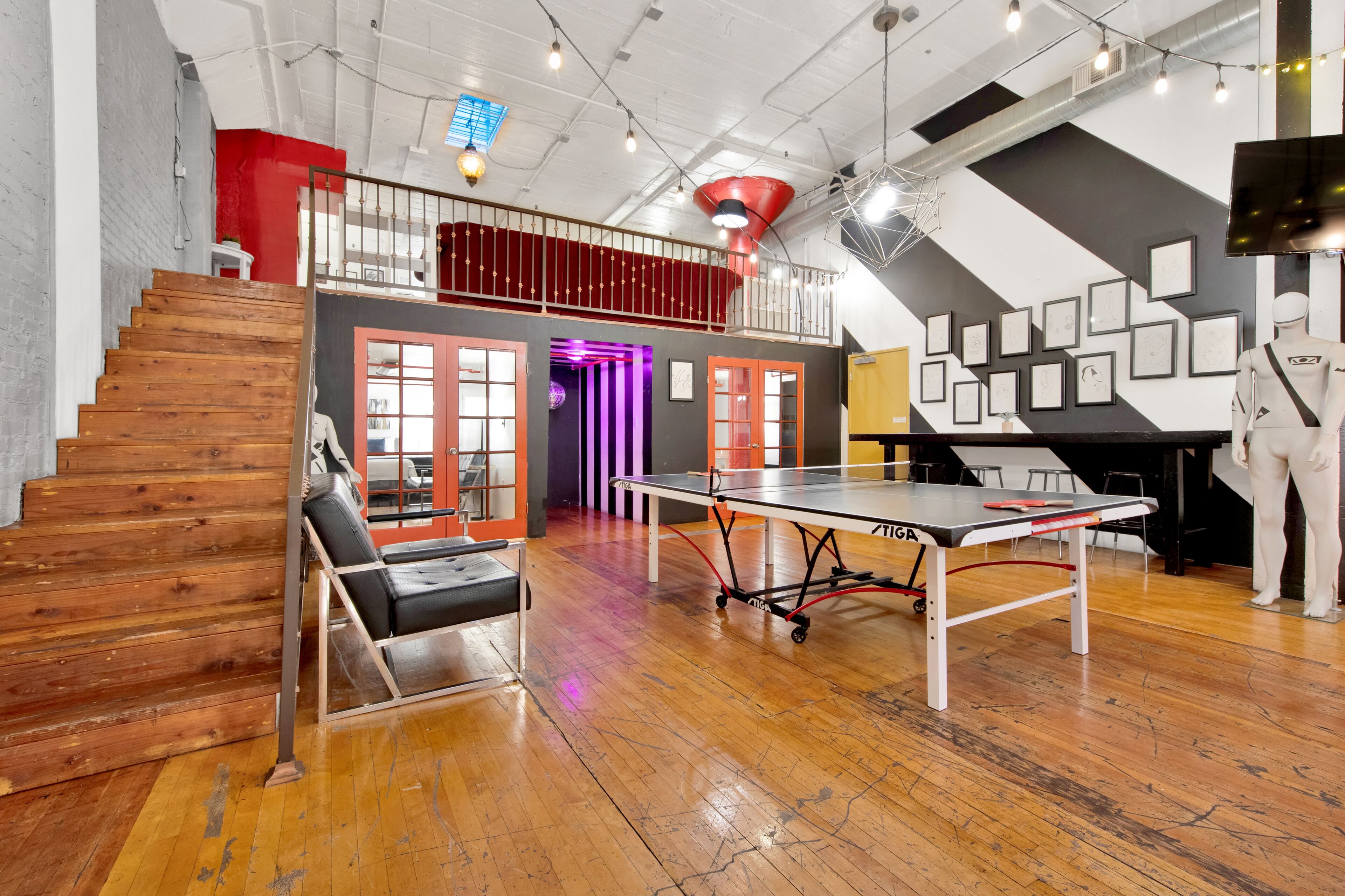 A spacious room with a ping pong table, wooden flooring, a staircase, and a mezzanine level with red railings.