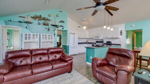 A spacious living room with a brown leather sofa, a wooden ceiling fan, and a kitchen area with white cabinets and a teal island. - Private Waterfront Retreat with Breathtaking Views. Pet-friendly! (Belhaven)