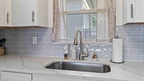 A kitchen with white cabinets, a stainless steel sink, a window with a curtain, and a tiled backsplash. - The Peaceful Place (Sarasota)