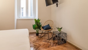 A room with a bed, a window, a television mounted on the wall, a wicker chair, and two potted plants. - Ghiberti Apartments - Persian Blue (Trieste)