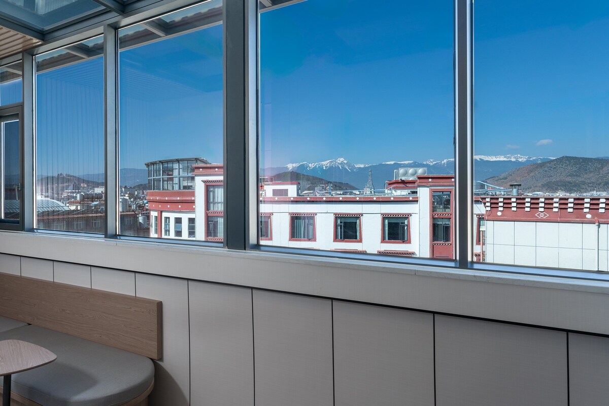 Een kamer met een groot raam, een bank en uitzicht op gebouwen en bergen.