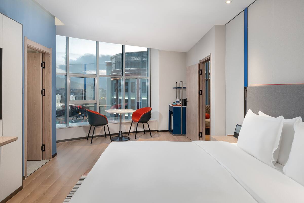 A modern hotel room with a large bed, a TV, a dining area with chairs, and a view of the city through large windows.