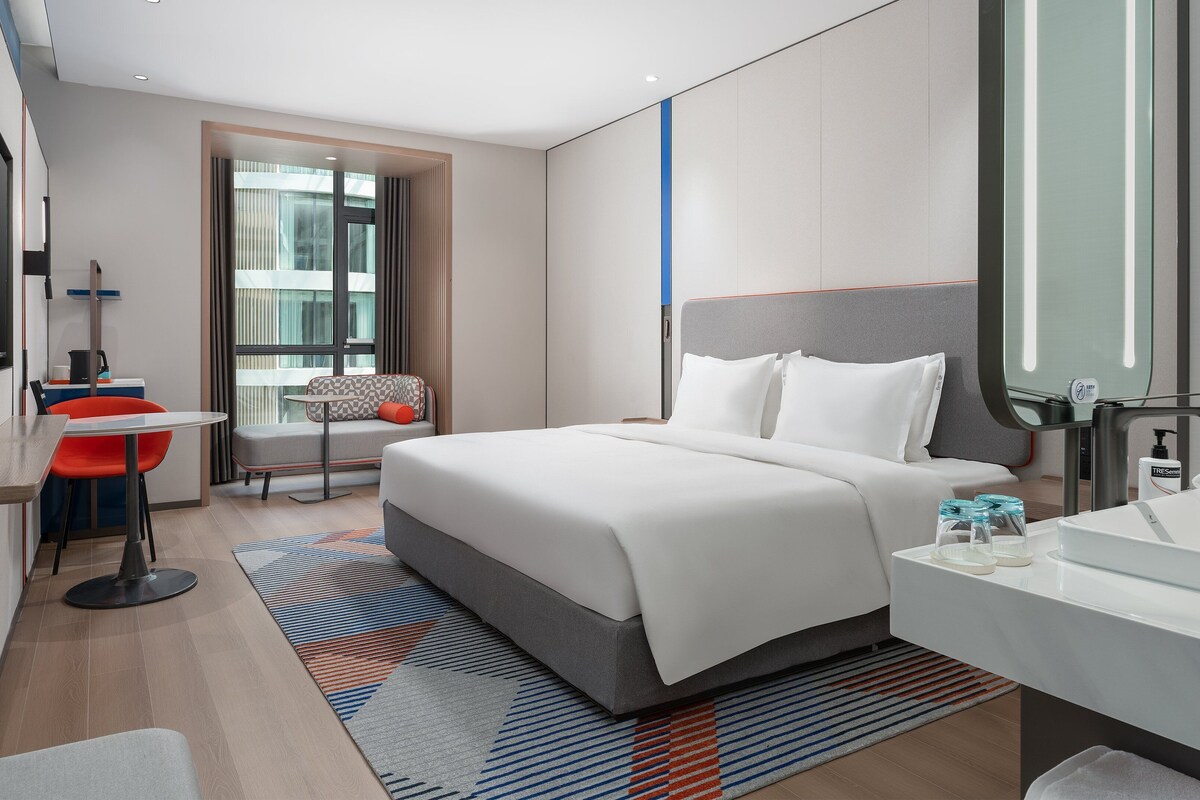 Een moderne hotelkamer met een groot bed, een zithoek met een stoel en tafel, een badkamer met wastafel en spiegel, en uitzicht op de stad door het raam.