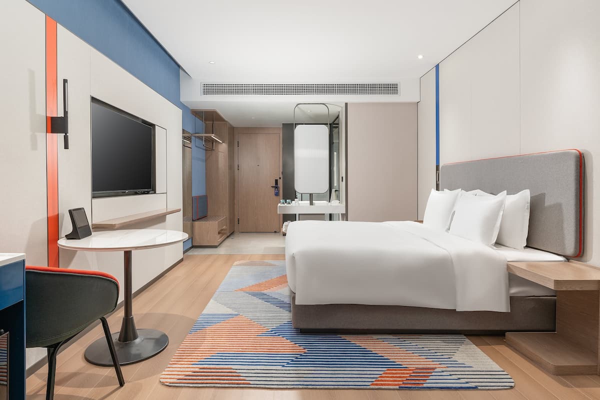 A modern hotel room with a bed, desk, chair, TV, and a bathroom visible through an open door.