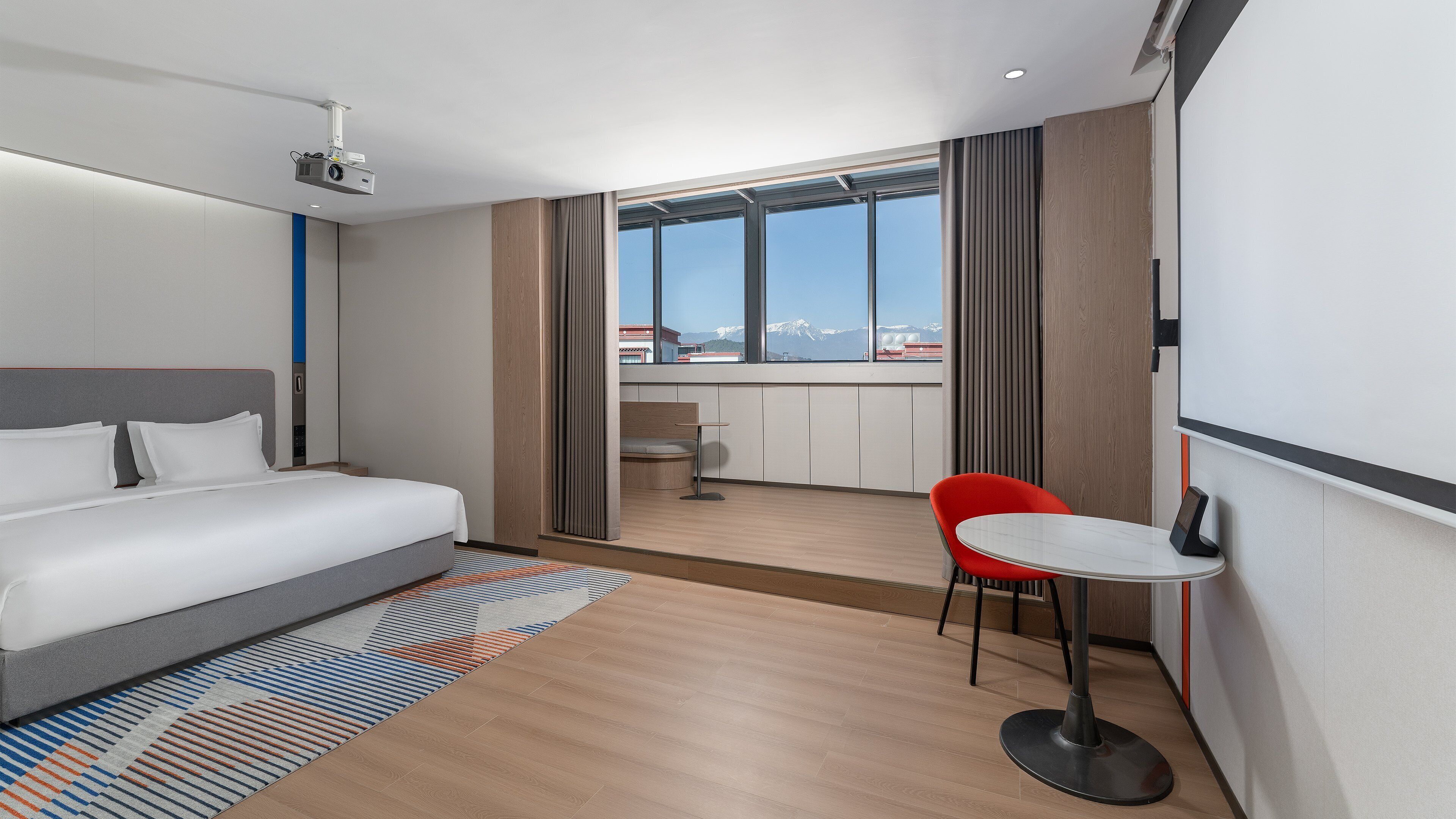 Ett modernt hotellrum med en säng, ett litet skrivbord, en röd stol och ett stort fönster med utsikt över staden.