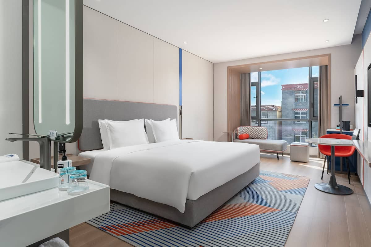 A modern hotel room with a large bed, a bathroom with a mirror and sink, a small seating area with a table, and a large window offering a city view.