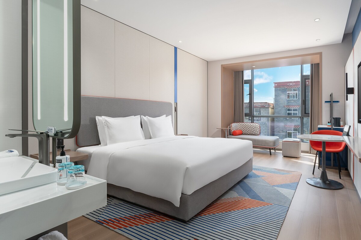 Een moderne hotelkamer met een groot bed, een badkamer met spiegel en wastafel, een klein zithoekje met een tafeltje en een groot raam met uitzicht op de stad.