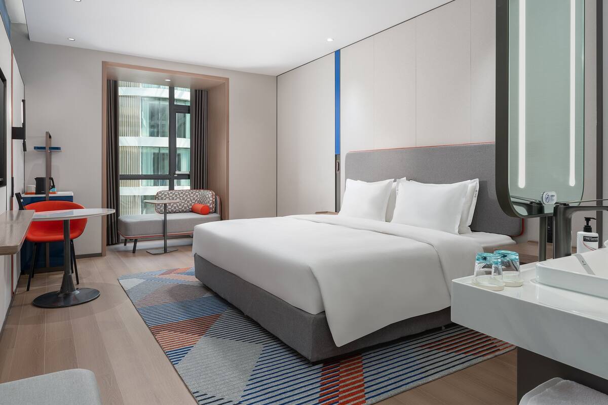 A modern hotel room with a large bed, a sitting area with a chair and table, a bathroom with a sink and mirror, and a view of the city through the window.