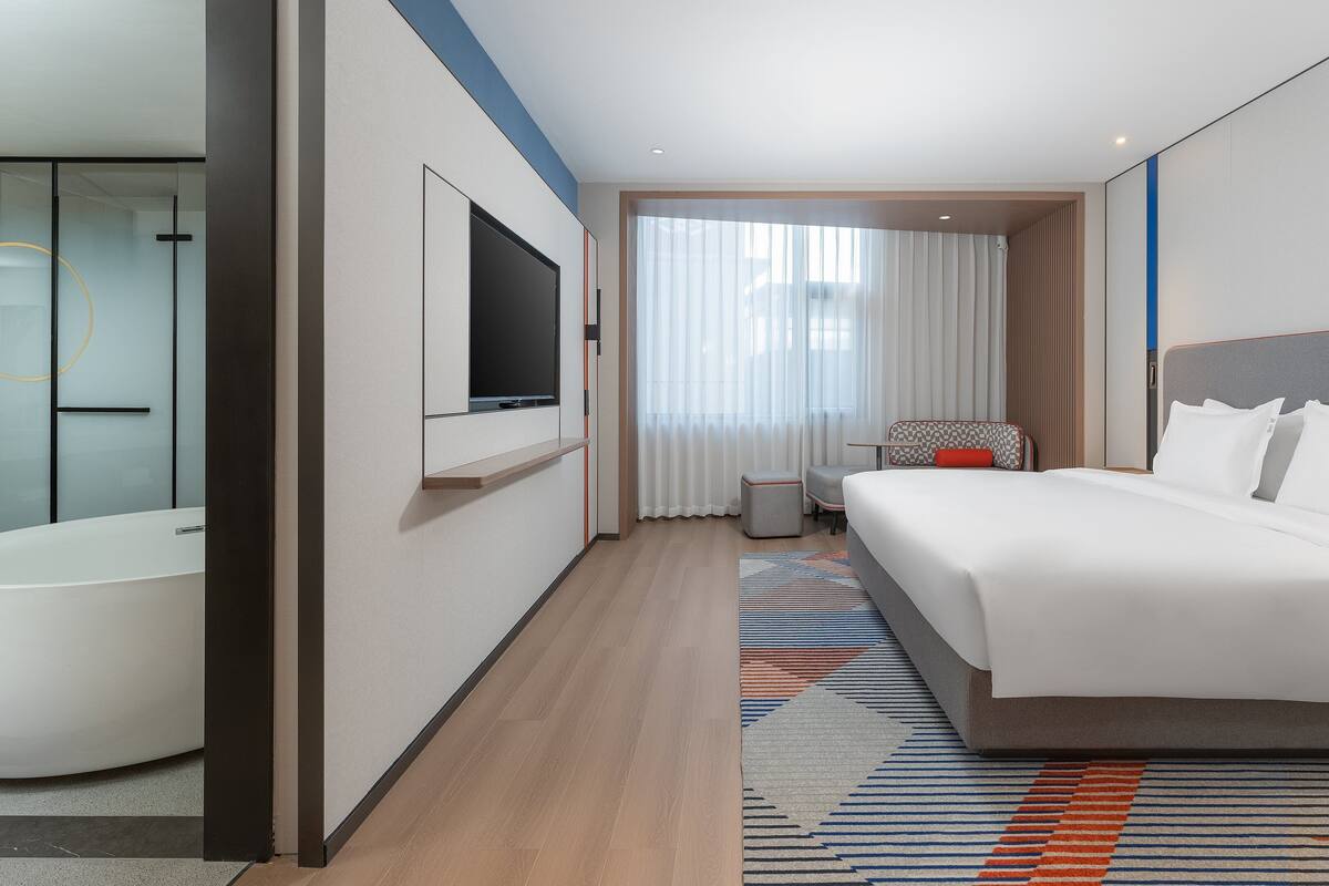 A modern hotel room with a large bed, a flat-screen TV, a separate bathroom with a bathtub, and a large window with sheer curtains.