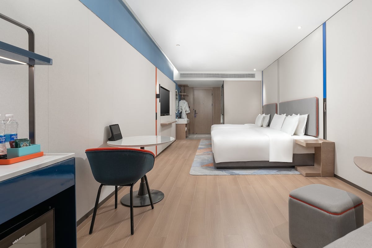 A modern hotel room with a bed, bedside table, chair, desk, and a small table.