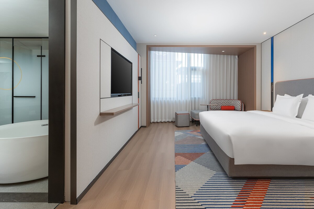 Een moderne hotelkamer met een groot bed, een flatscreen televisie, een badkamer met badkuip en een groot raam met gordijnen.