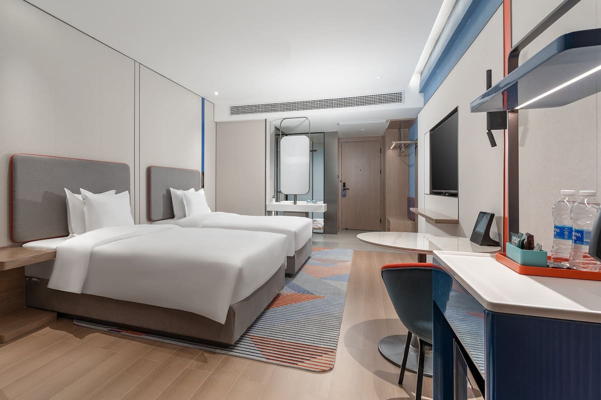 A modern hotel room with a large bed, a desk with a chair, a mirror, and a television.