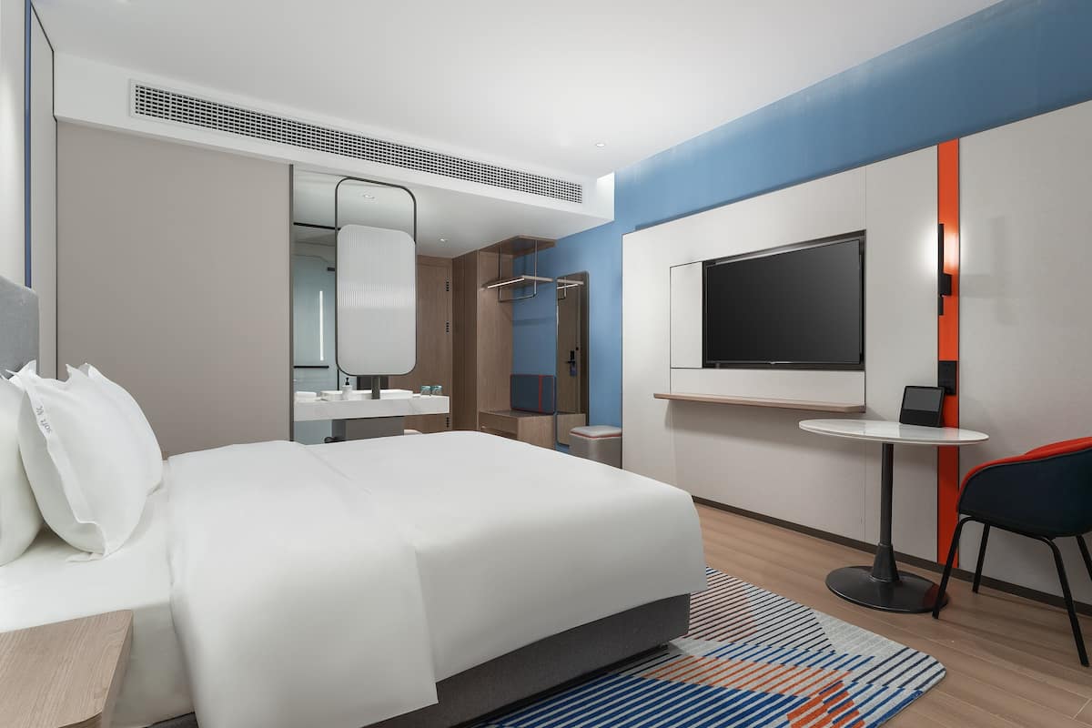 A modern hotel room with a large bed, a desk, a chair, a TV, and a minibar.