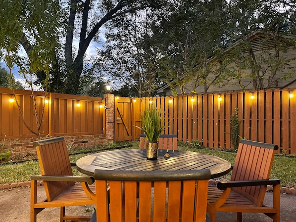 An outdoor patio with a round table and four wooden chairs, surrounded by a wooden fence and a brick wall.