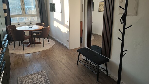 Een moderne woonkamer met een ronde eettafel, twee stoelen, een bankje, een kapstok en een groot raam met gordijnen.