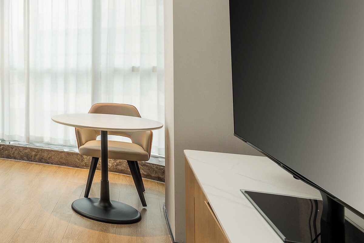 A modern room with a round table and a single chair, a large flat-screen TV mounted on the wall, and a window with sheer curtains.