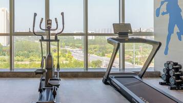 A gym with a treadmill, elliptical, and dumbbells, with a city view through large windows.