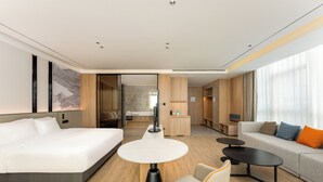A modern hotel room with a bed, a round table, a sofa, and a small kitchen area.