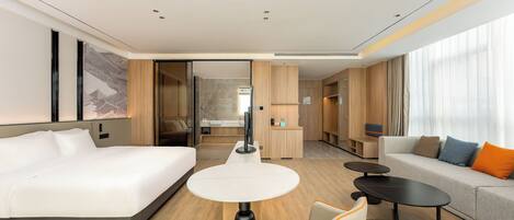 A modern hotel room with a bed, a round table, a sofa, and a small kitchen area.