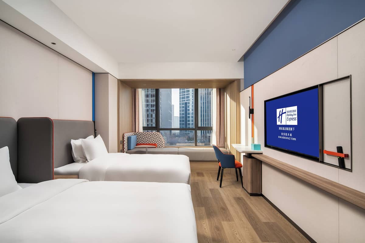 A hotel room with two beds, a desk, a chair, and a TV screen displaying hotel information.