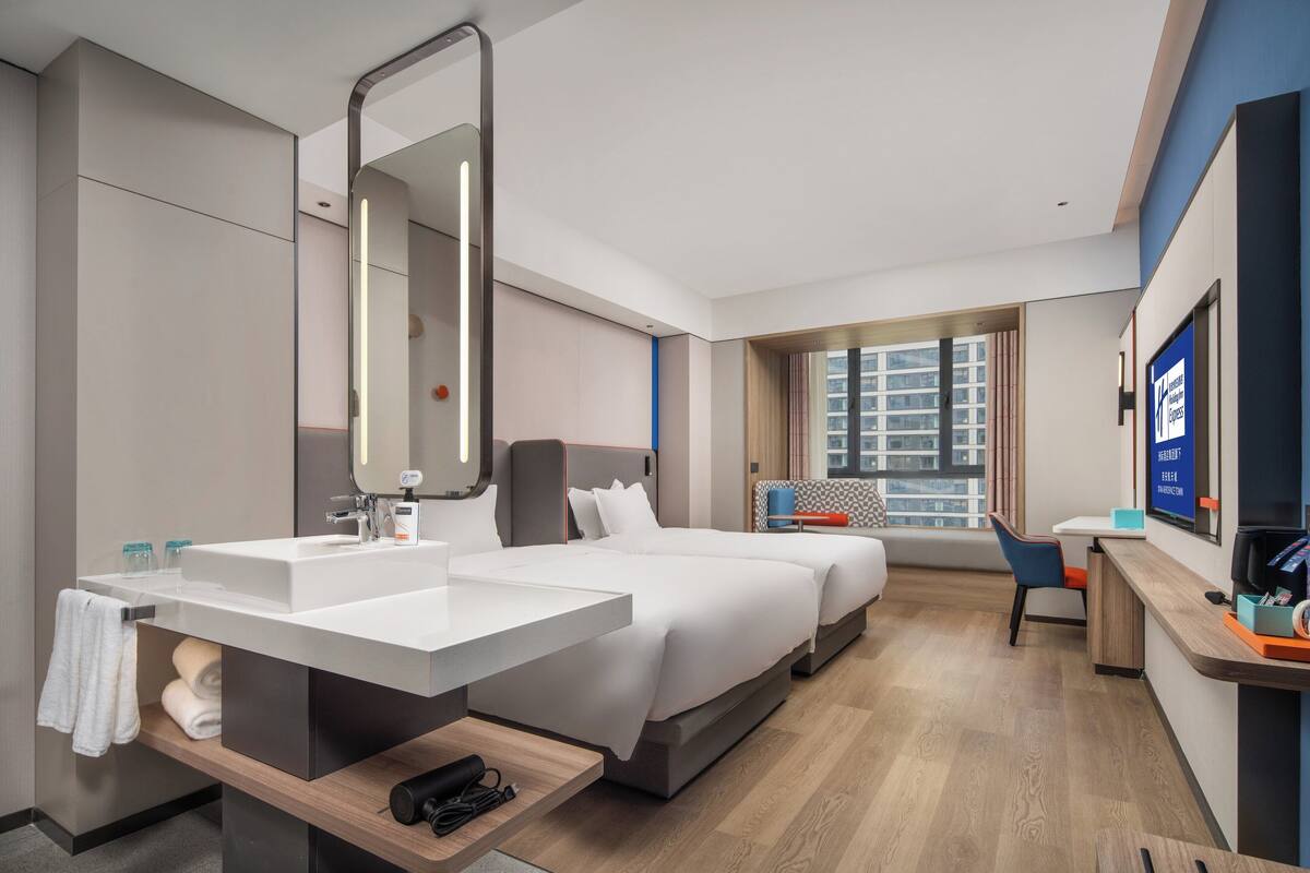 A modern hotel room with a large bed, a desk, a bathroom with a sink, and a large window offering a city view.