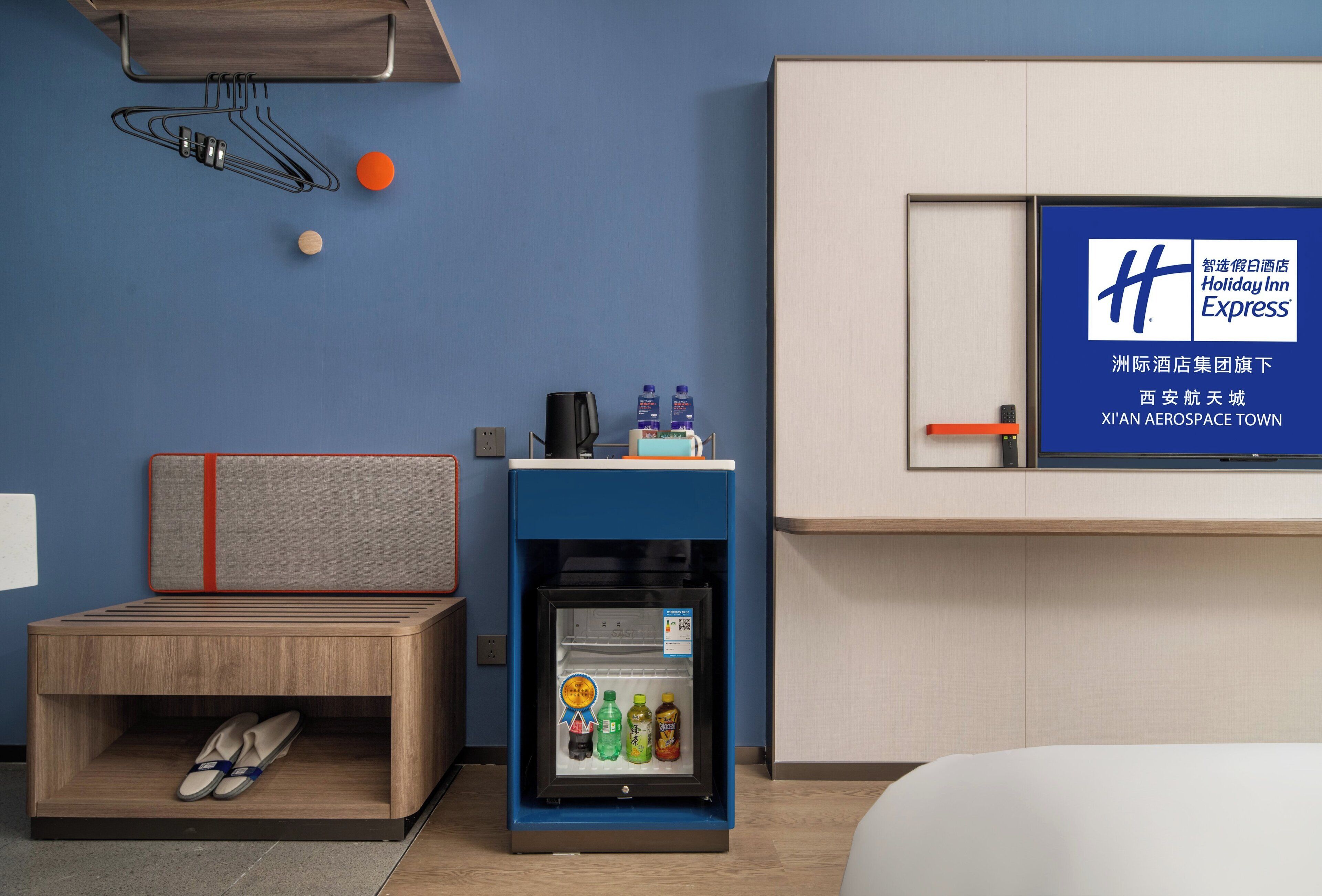 Une chambre d’hôtel comprenant un lit, une petite table de chevet en bois avec des chaussons, un mini-réfrigérateur contenant des boissons et un téléviseur fixé au mur affichant le logo de Holiday Inn Express.