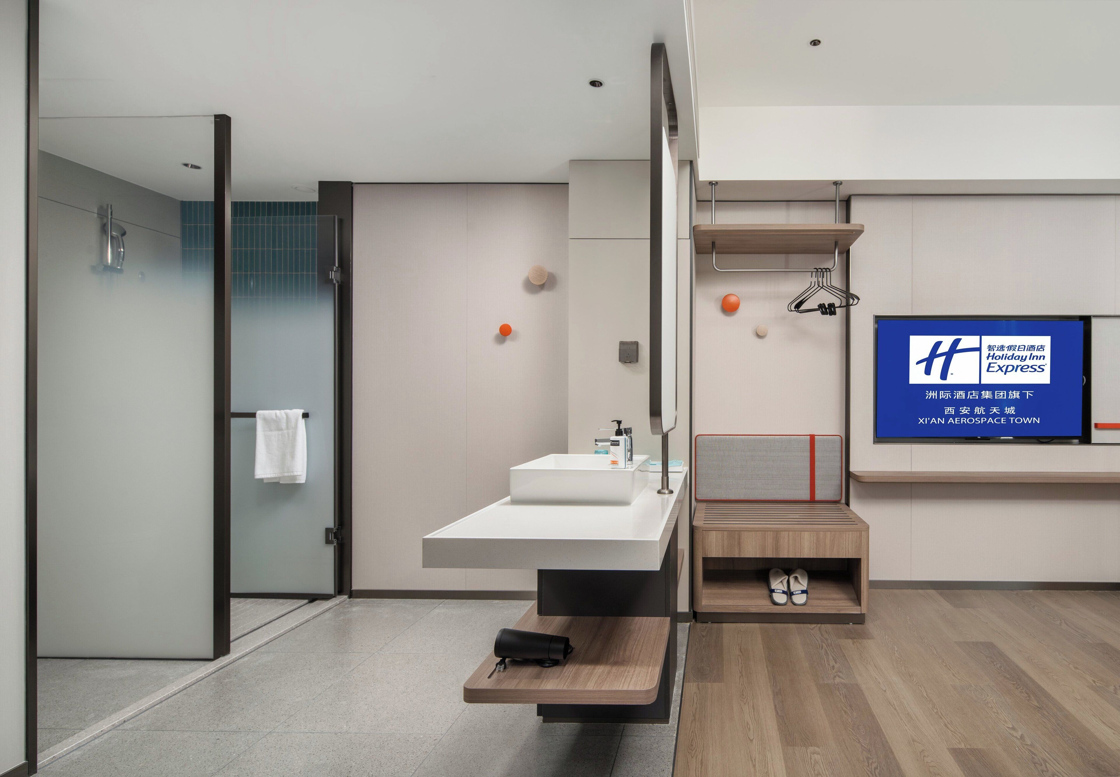 A modern hotel bathroom with a large sink, a mirror, and a television displaying the Holiday Inn Express logo.