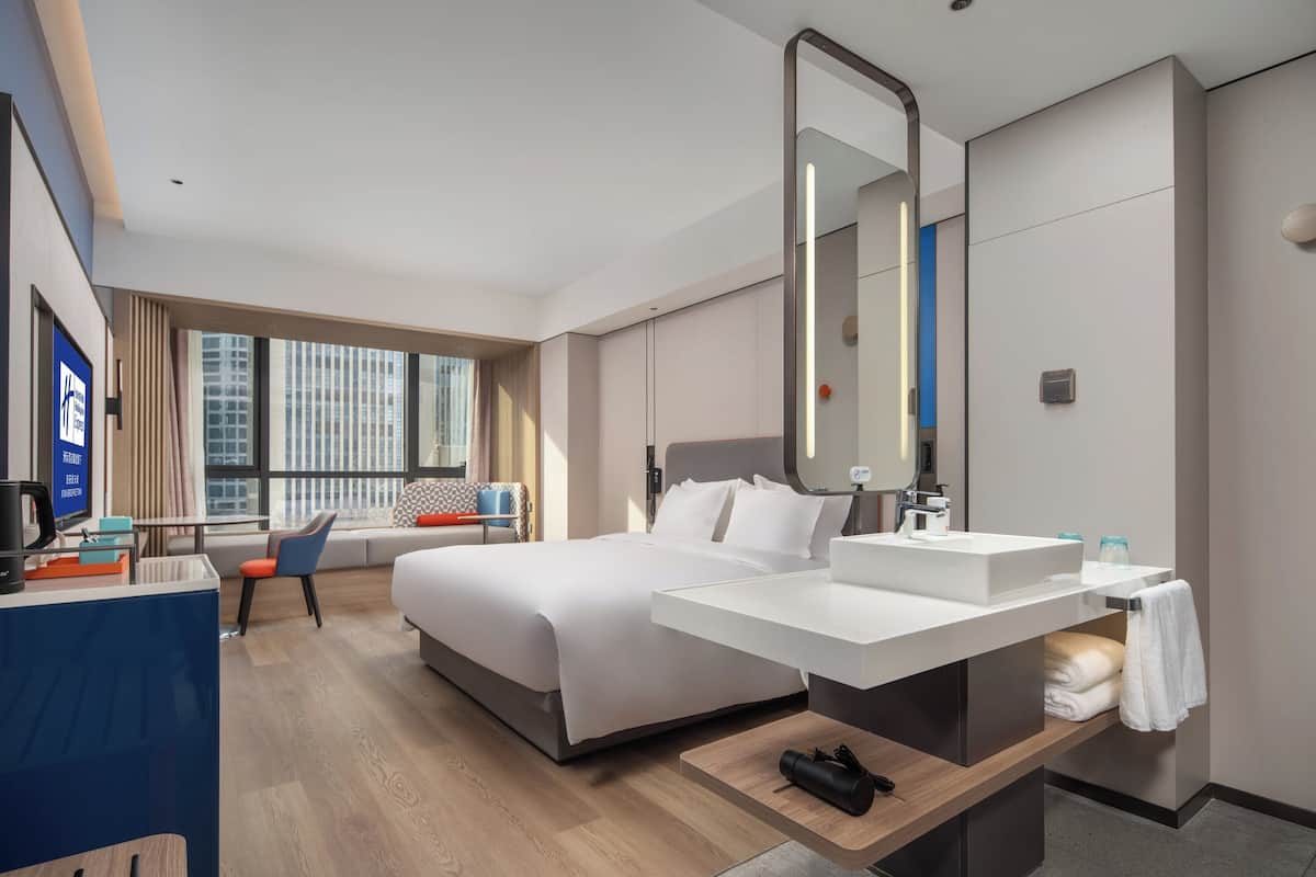 A modern hotel room with a large bed, a desk, a bathroom with a large mirror, and a view of the city through large windows.