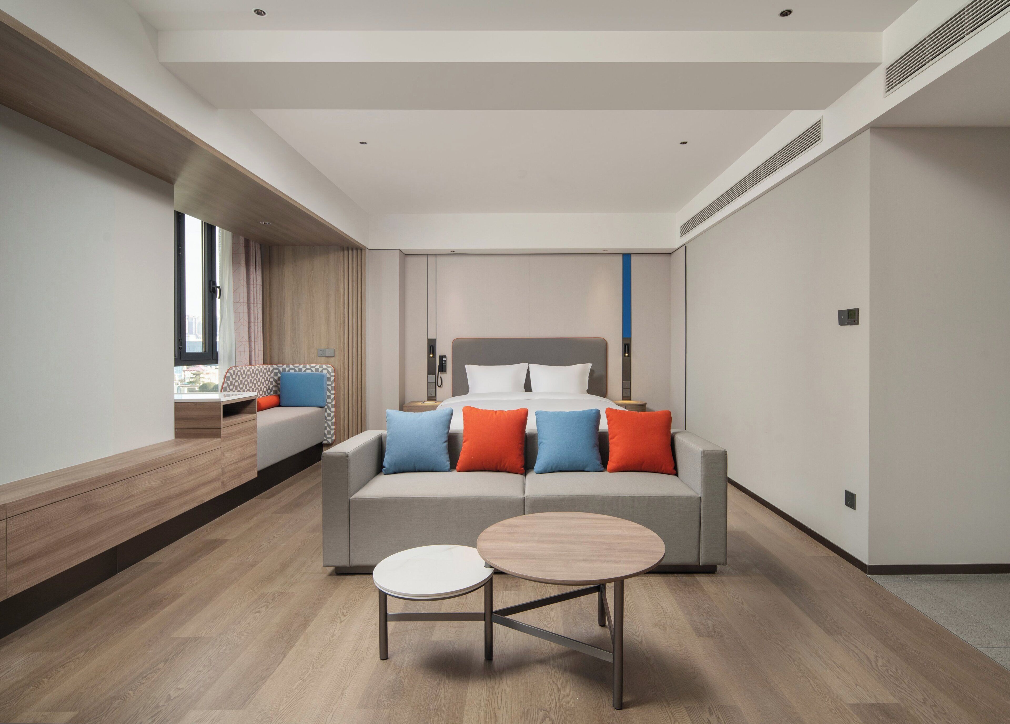 A modern hotel room with a large bed, a sofa, a small table, and a window with curtains.