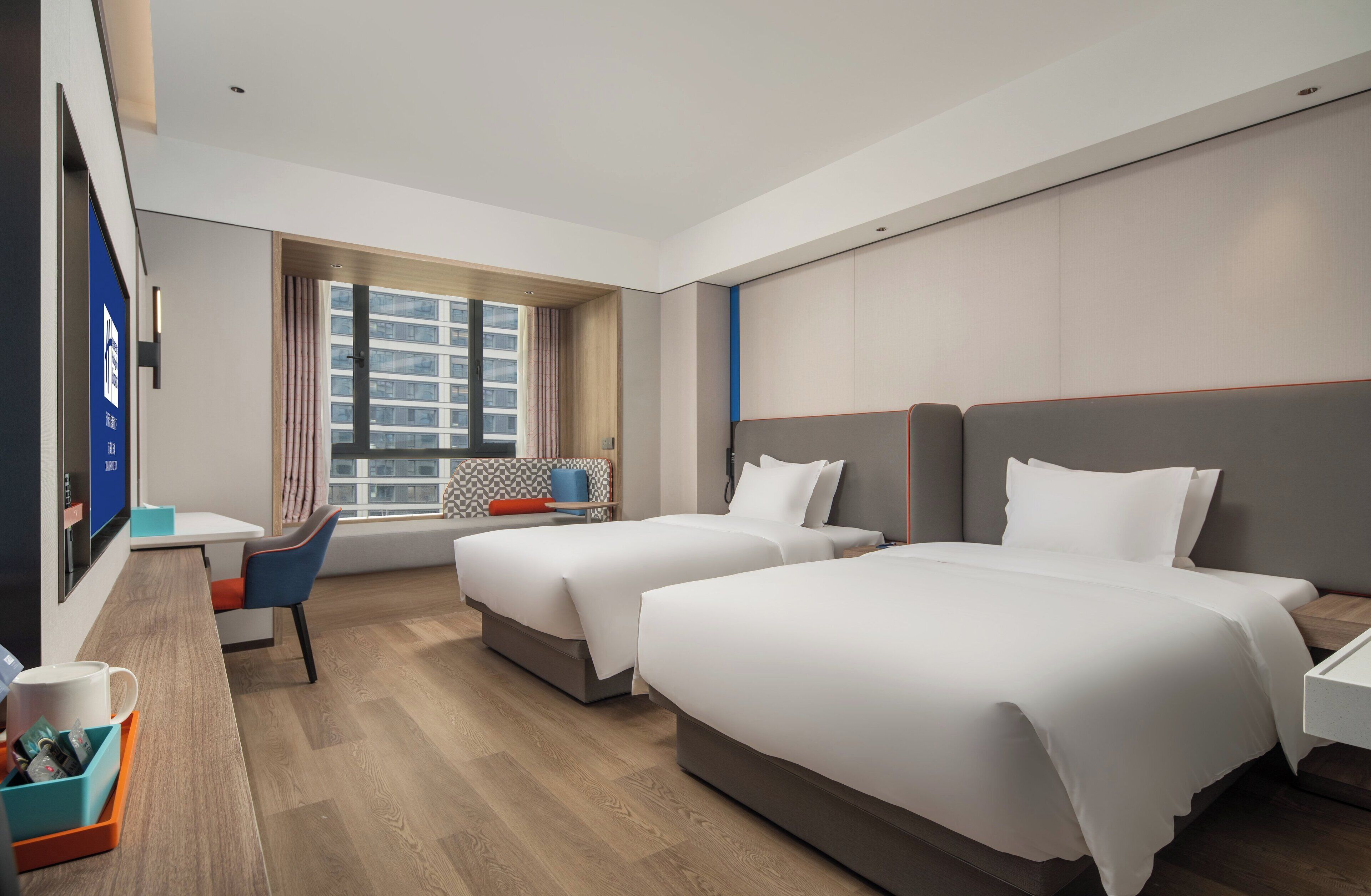 Ein Hotelzimmer mit zwei Betten, einem Schreibtisch, einem Stuhl, einem Fenster mit Vorhängen und Blick auf Gebäude.