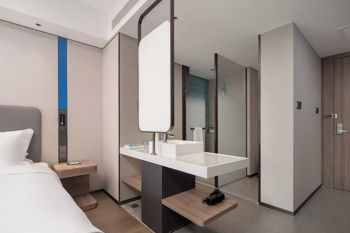 A modern hotel room with a large bed, a white vanity with a mirror, a wooden nightstand, and a bathroom visible through a glass door.