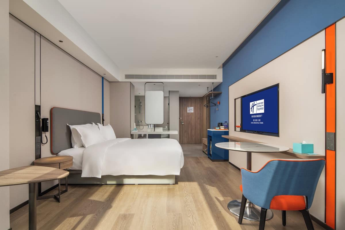 A modern hotel room with a bed, bedside table, chair, TV, and a desk with a chair.