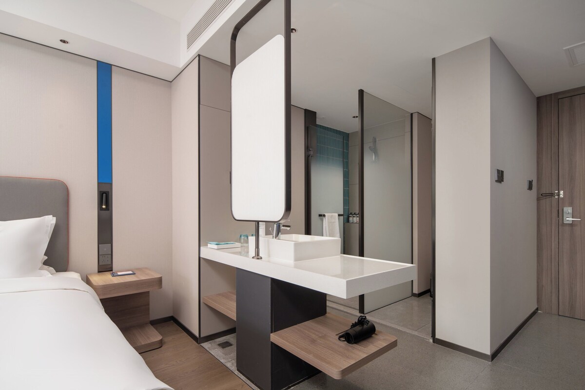 A modern hotel room with a large bed, a white vanity with a mirror, a wooden nightstand, and a bathroom visible through a glass door.