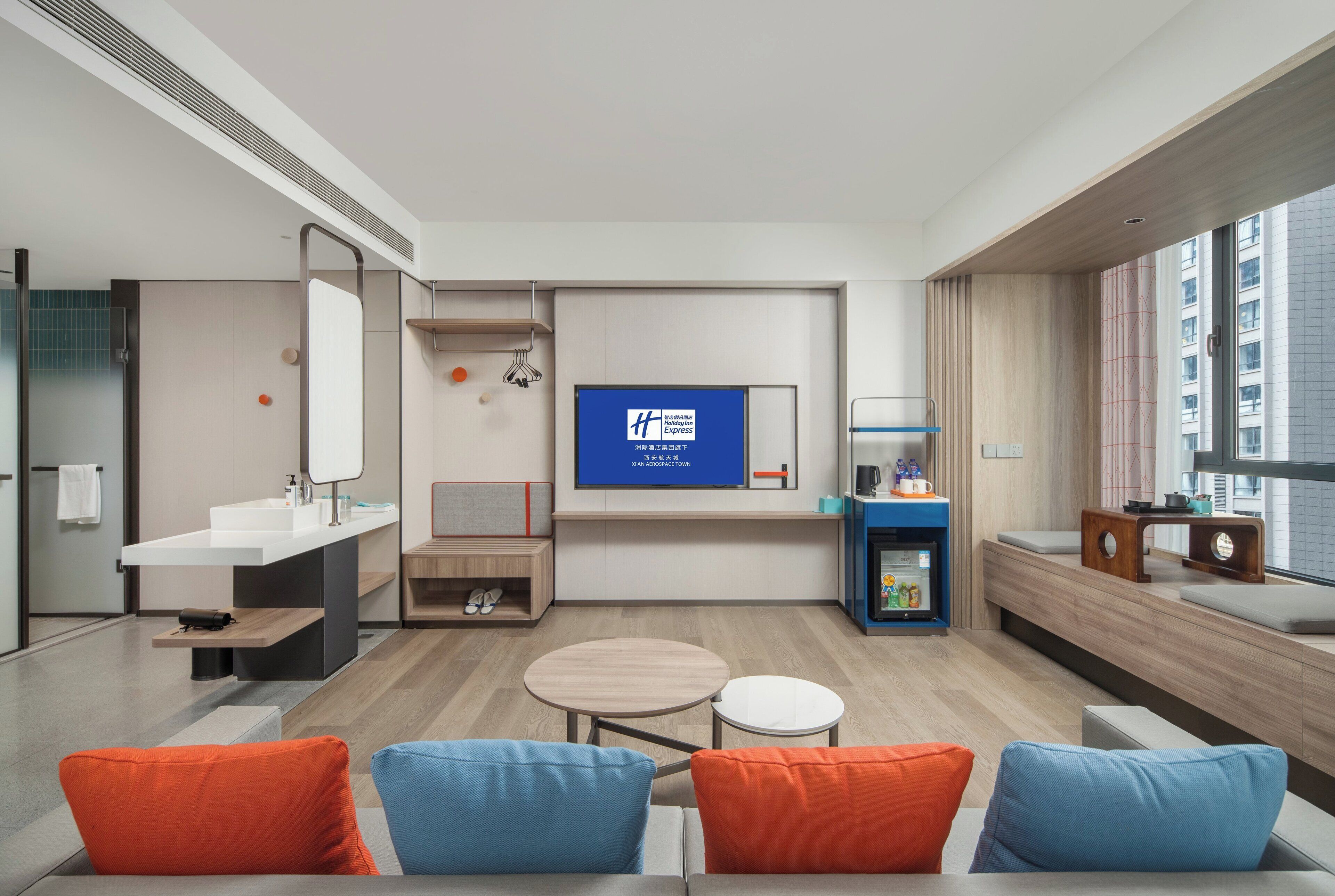 A modern hotel room with a large TV, a sofa with colorful cushions, a small table, a built-in bench, and a large window offering a city view.