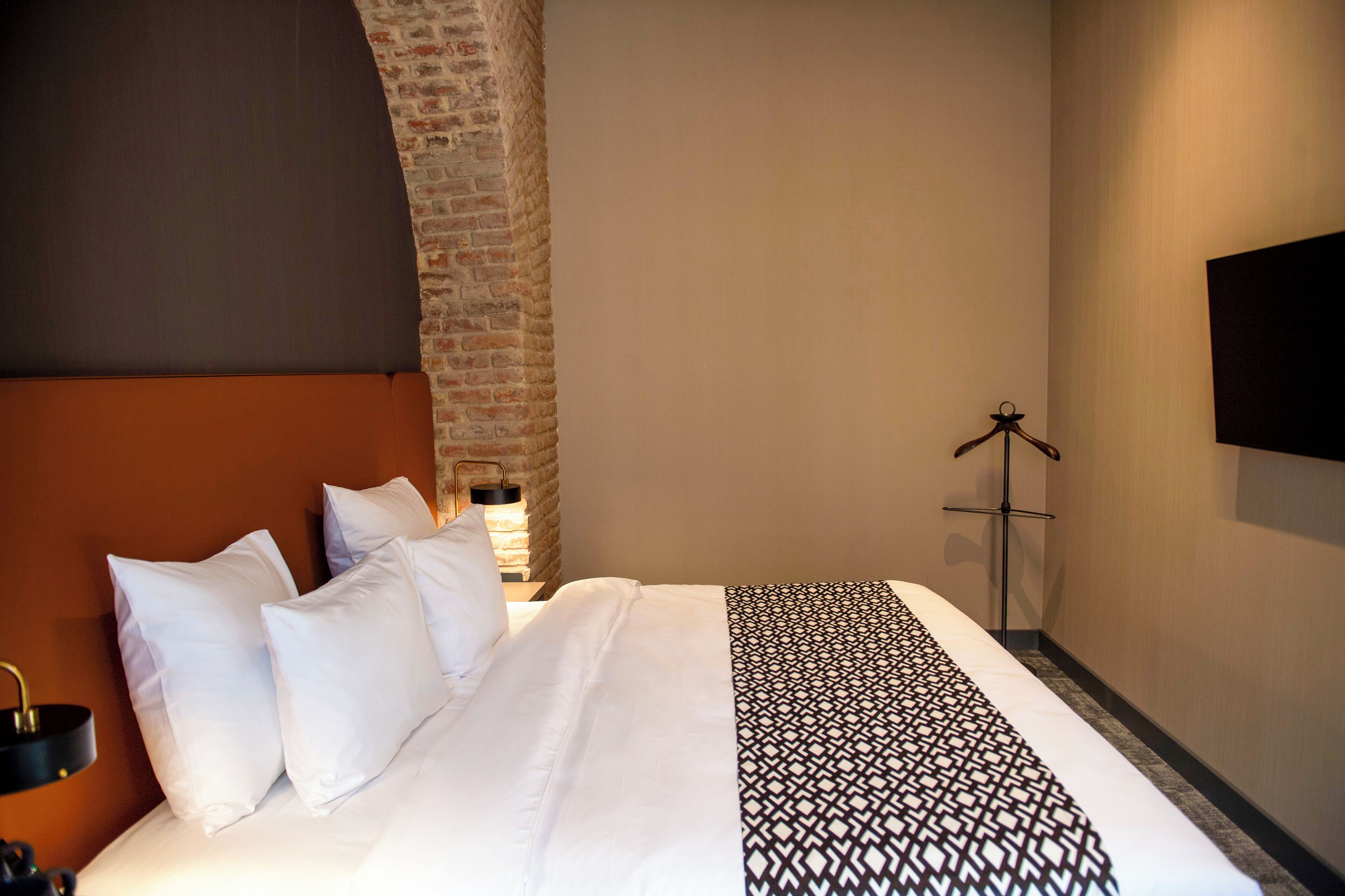 Habitación de hotel con pared de ladrillo, cama con ropa de cama blanca, manta estampada y televisor montado.