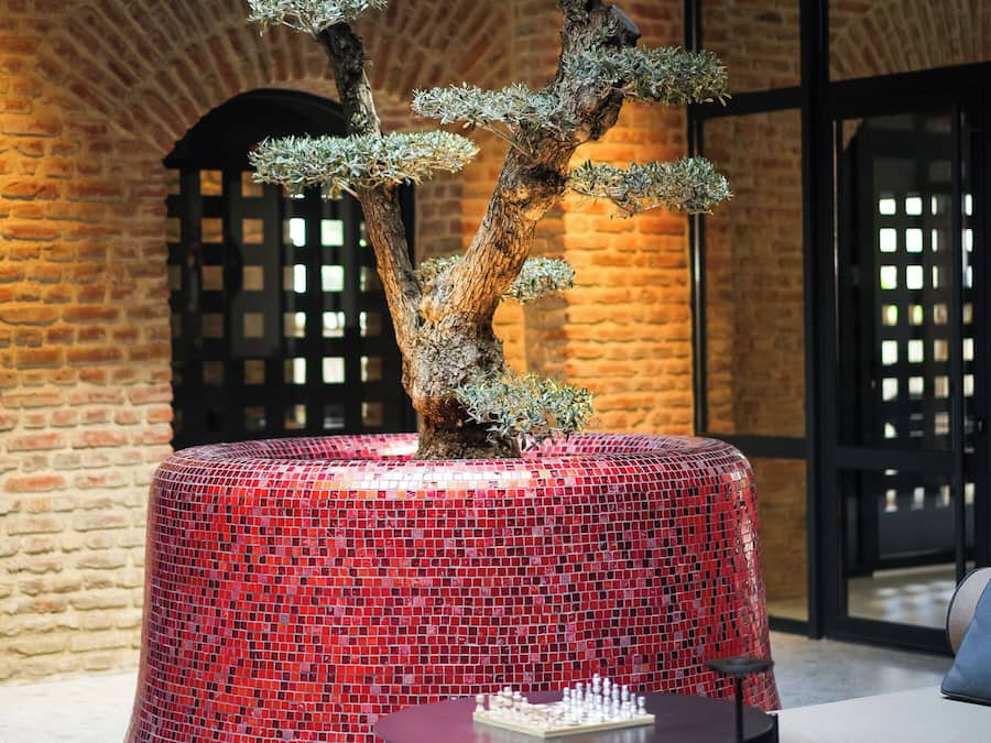 A bonsai tree on a red mosaic pedestal in a modern interior with brick walls and a chess table.