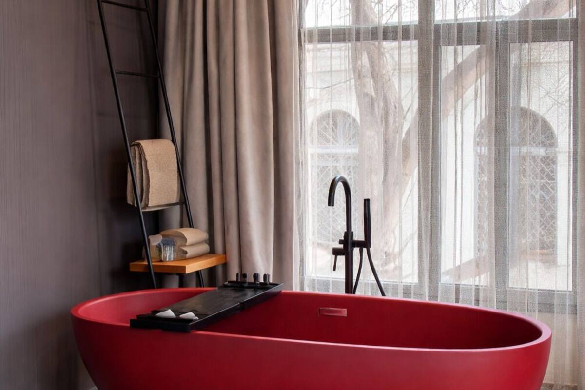 Un baño moderno con una bañera independiente color rojo, un estante de madera con toallas y una ventana con cortinas traslúcidas.