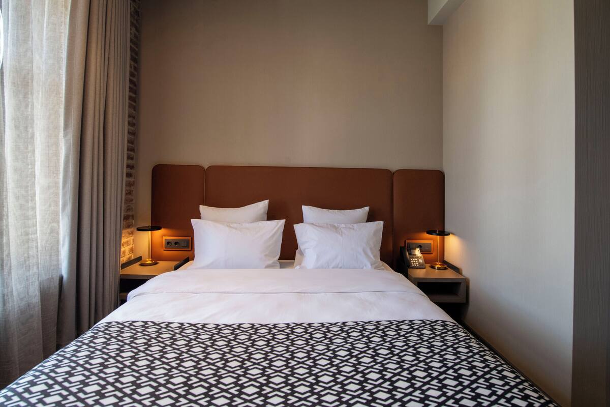 Una habitación de hotel con una cama, dos mesitas de noche, una lámpara y una cortina.