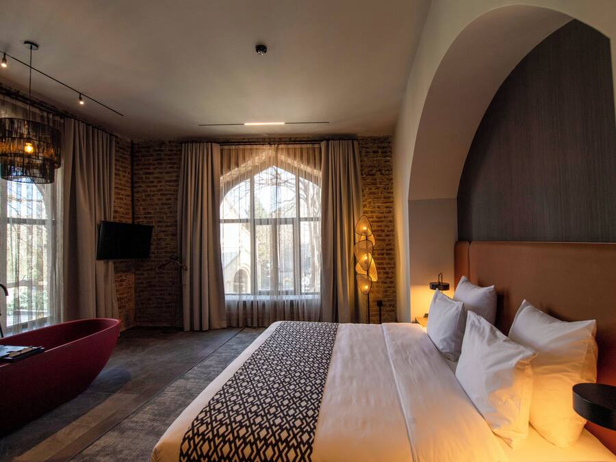 Quarto de hotel com uma cama grande, uma banheira vermelha, uma televisão e uma janela com cortinas.