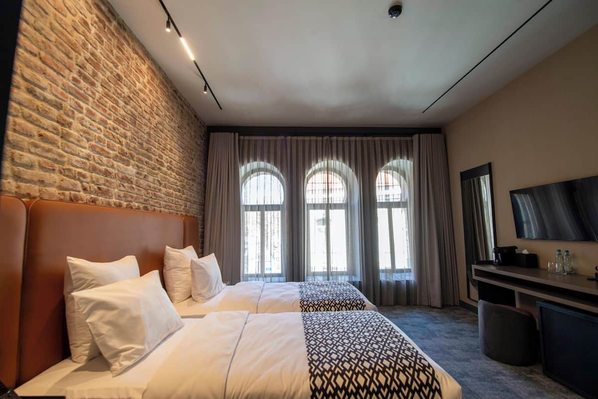 Habitación de hotel con pared de ladrillo, dos camas con ropa blanca, un televisor grande y un escritorio con espejo y botellas.