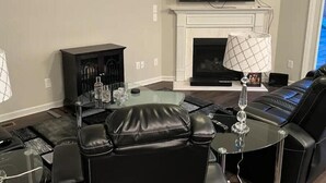 A living room with a black leather sofa, a glass coffee table, a fireplace, a flat-screen TV, and framed pictures on the wall.