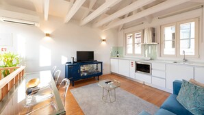 A modern living room with a glass dining table, a blue sofa, and a kitchen with white cabinets and a built-in oven.