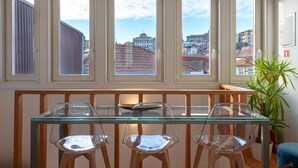 A dining area with a glass table and three chairs, large windows with a city view, and a potted plant.