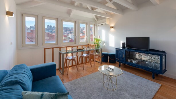 A spacious living area with a blue sofa, a glass dining table with chairs, a TV on a blue cabinet, and large windows with a city view.