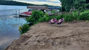 A lakeside view with a sandy beach, two folding chairs, a dock with boats, and a covered area.