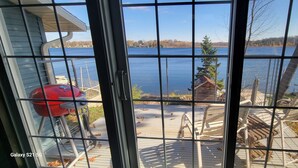 A view through a window with a grill and chairs on a deck overlooking a lake.