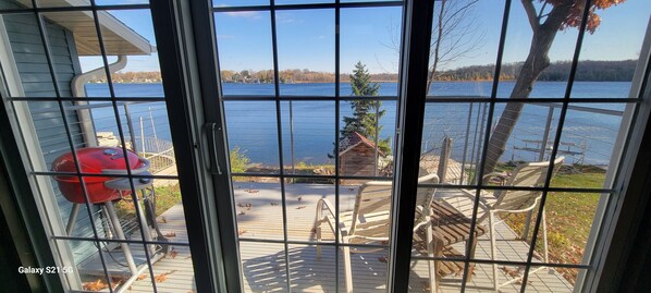Vista a través de una ventana con una parrilla y sillas en una terraza con vista a un lago.