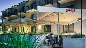 Un área de estar al aire libre con mesas y sillas bajo sombrillas, un edificio con balcones y un letrero que dice "ŁŻWIEC".